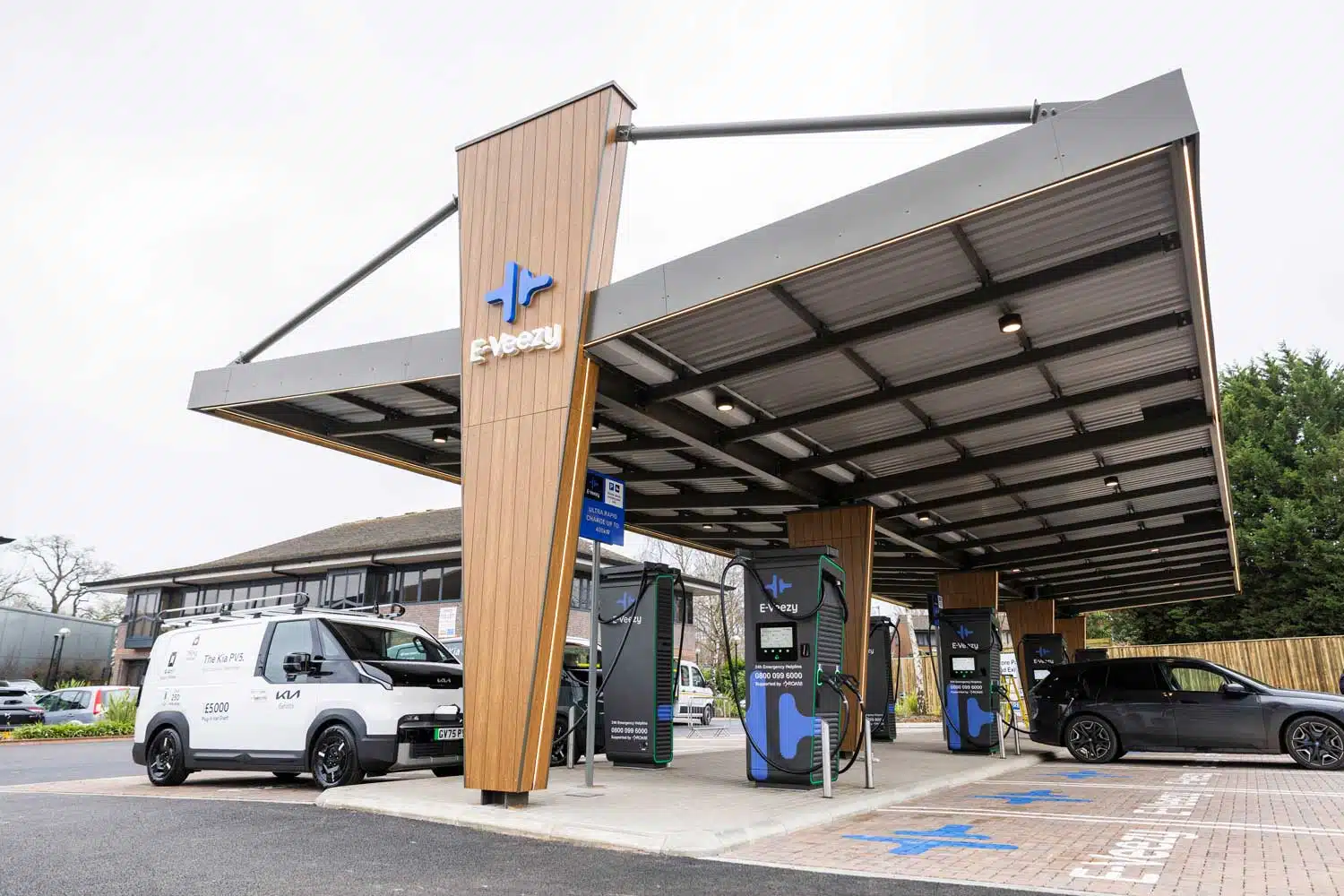 Person charging electric vehicle at branded E‑Veezy station in landscaped commercial setting with canopy and fleet vehicles.