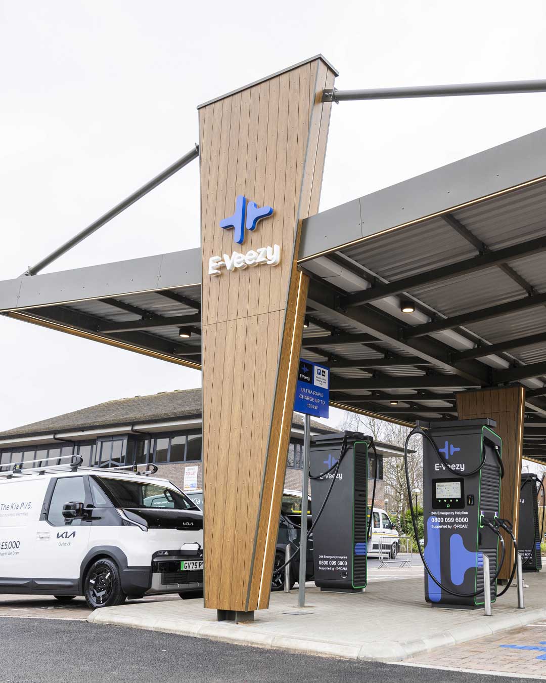 Person charging electric vehicle at branded E‑Veezy station in landscaped commercial setting with canopy and fleet vehicles.