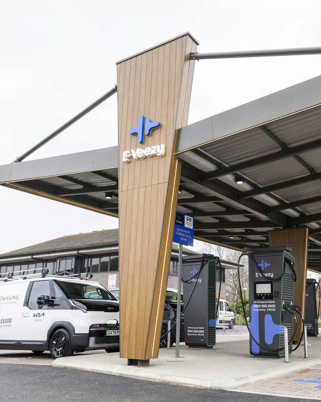 Person charging electric vehicle at branded E‑Veezy station in landscaped commercial setting with canopy and fleet vehicles.
